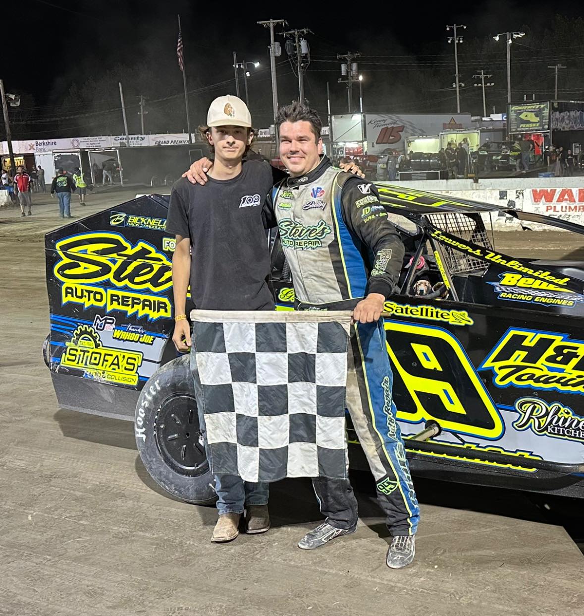 Kolby and Christopher holding checkered flag with Car #99 behind them in victory lane on dirt at night